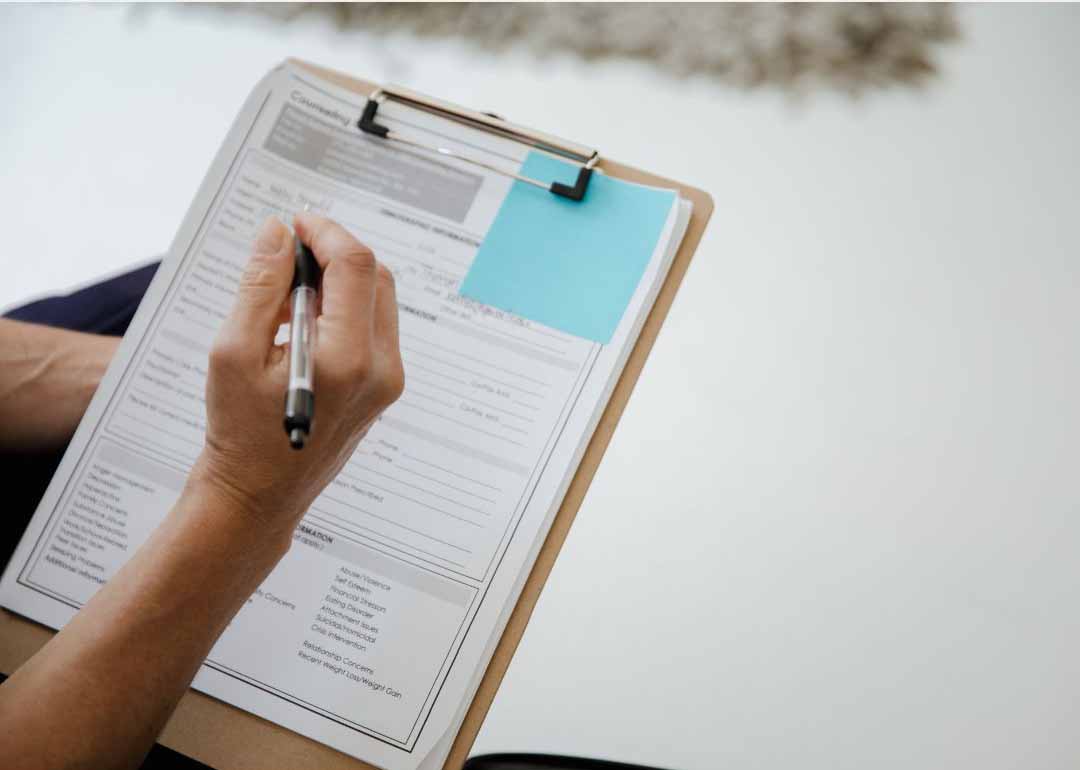 Person's hand writes with a pen on a form clipped to a clipboard, with a blue sticky note attached.